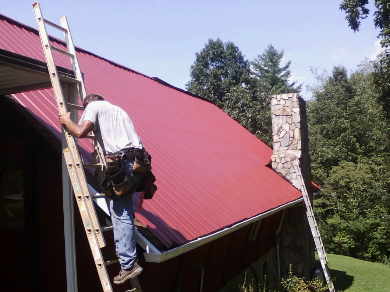 Roofer on a ladder beside a red metal roof and stone chimney — Soffit & Fascia Repair by Roofing Brothers in Council
